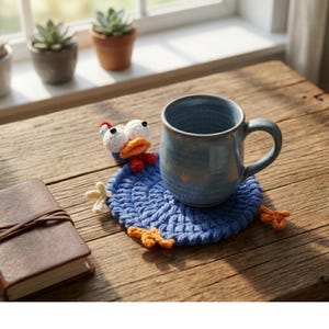 May include: A blue ceramic mug rests on a handmade blue crochet coaster with a whimsical bird design. The coaster features white eyes, an orange beak, and yellow feet. The mug and coaster sit on a wooden table.