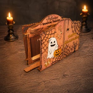 May include: A wooden Halloween Shadow Theatre with laser-cut details including a ghost holding a pumpkin, spider webs, and trees. The front panels feature the text 'Halloween SHADOW THEATRE' and open to reveal a stage area with curtain designs.