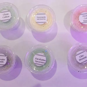 Könnte beinhalten: Sechs kleine, transparente Plastikbehälter gefüllt mit Duftwachs-Schmelzen. Jede Schmelze hat eine andere Farbe und einen anderen Duft, beschriftet mit Text wie "Clean Cotton", "Honeysuckle and Jasmine" und "Fresh Cut Roses". Die Schmelzen sind auf einer hellvioletten Oberfläche angeordnet.