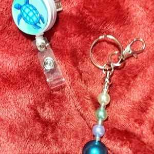 May include: A white retractable badge reel featuring a blue sea turtle graphic and a clear plastic strap, alongside a keychain with a silver metal ring and clasp, decorated with a vertical row of four graduated round beads in cream, mint, lavender, and blue.