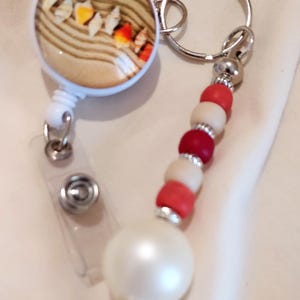 Può includere: Un portabadge con un inserto circolare raffigurante una spiaggia sabbiosa e conchiglie, fissato a un portachiavi in metallo con un cordino di perline. Il cordino è composto da perline rosse e color crema alternate, distanziatori argentati e una grande perla bianca.