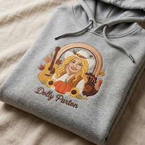 May include: A heather gray hooded sweatshirt featuring an embroidered design of a blonde person in a cowboy hat, a guitar, a pumpkin, sunflowers, autumn leaves, and a turkey. The text Dolly Parton is embroidered in brown below the design.