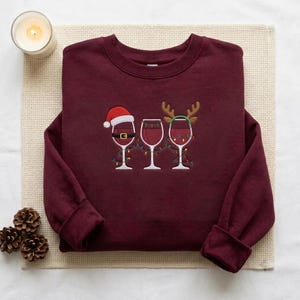 May include: A maroon crewneck sweatshirt featuring an embroidered graphic of three wine glasses filled with red wine. The first glass wears a Santa hat and belt, the second is plain, and the third wears reindeer antlers, all connected by string lights.