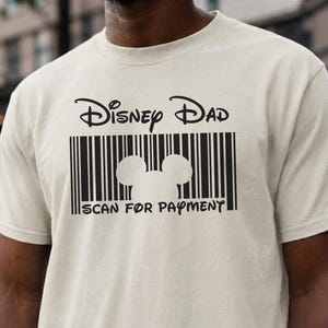 以下が含まれることがあります： ナチュラルカラーのTシャツに黒のグラフィックプリントが施されています。デザインには「Disney Dad」という文字、中央にミッキーマウスの頭のシルエットがあるバーコード、そしてバーコードの下に「SCAN FOR PAYMENT」という文字が含まれています。