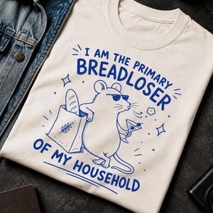 May include: An ivory t-shirt featuring a blue graphic of a mouse wearing sunglasses, holding a martini glass, and standing next to a paper bag of bread. The text reads, I AM THE PRIMARY BREADLOSER OF MY HOUSEHOLD, printed in blue ink.