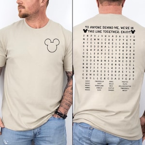 May include: A beige t-shirt featuring a black Mickey Mouse head outline on the front left chest. The back displays a word search puzzle with the text "TO ANYONE BEHIND ME, WE'RE IN THIS LINE TOGETHER. ENJOY!" and a list of Disney-themed words below.