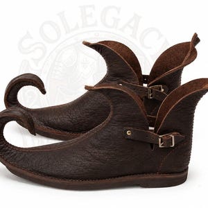 May include: A pair of dark brown leather shoes featuring a distinctively elongated, upward-curving toe and a high, flared ankle collar. The shoes are secured with a thin leather strap and a small metal buckle on the side, showcasing a textured finish.
