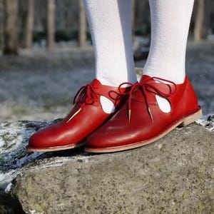 Puede incluir: Zapatos de cuero rojo con un diseño calado único y cordones rojos a juego. Los zapatos se usan con calcetines blancos. Los zapatos están sobre una roca gris, con un fondo borroso de árboles.