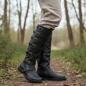 May include: A pair of knee-high black boots featuring a textured finish and multiple horizontal straps secured with antique brass-toned metal buckles running up the front of the shaft, paired with light beige trousers tucked into the boots.