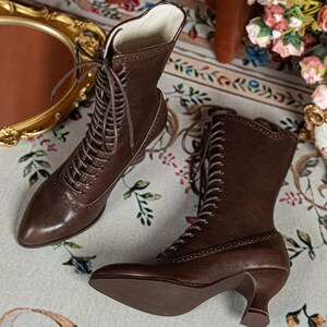 K&ouml;nnte beinhalten: Ein Paar wadenhohe Stiefel aus dunkelbraunem Leder mit spitzer Zehenpartie, Schn&uuml;rung vorne mit passenden braunen Schn&uuml;rsenkeln und einem geschwungenen Kitten-Heel-Absatz. Die Stiefel weisen dekorative Perforationen im Brogue-Stil entlang der N&auml;hte auf.