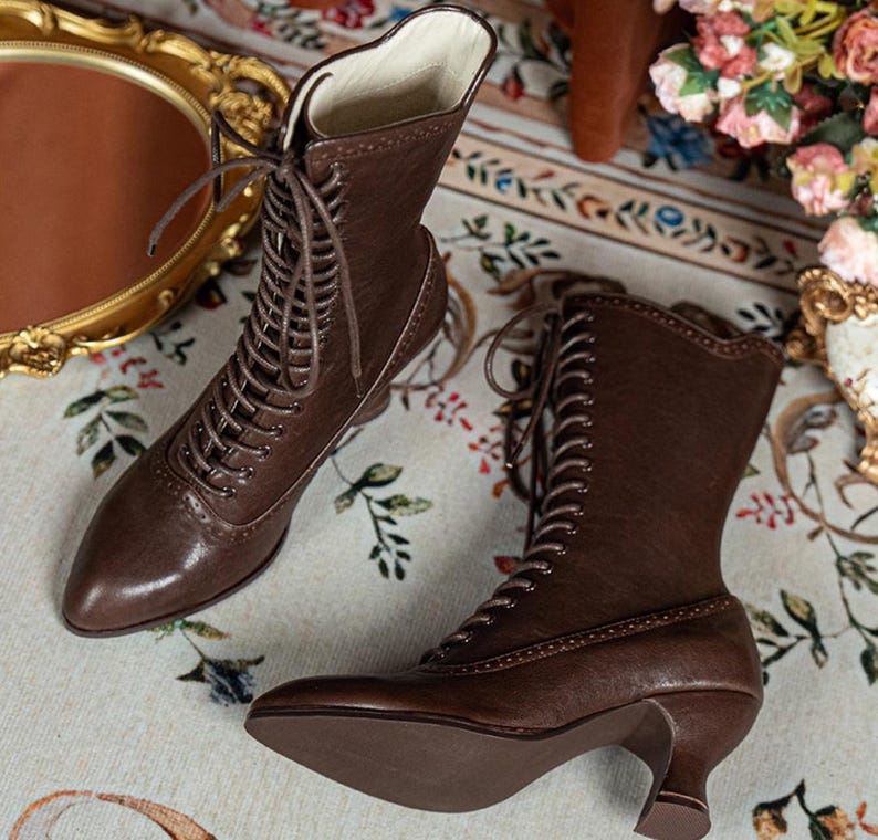 K&ouml;nnte beinhalten: Ein Paar wadenhohe Stiefel aus dunkelbraunem Leder mit spitzer Zehenpartie, Schn&uuml;rung vorne mit passenden braunen Schn&uuml;rsenkeln und einem geschwungenen Kitten-Heel-Absatz. Die Stiefel weisen dekorative Perforationen im Brogue-Stil entlang der N&auml;hte auf.