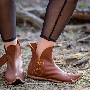 May include: A pair of brown leather ankle boots featuring a pointed toe, a high-cut curved collar, and side lacing with thin orange laces. The boots have visible stitching along the seams and a flat sole, designed with a distinct tapered silhouette.