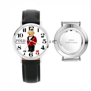 May include: A wristwatch with a black leather strap and a white face featuring a graphic of a teddy bear in a red tuxedo, with the text POLO RALPH LAUREN. Beside it is a separate silver-toned watch case back engraved with POLO RALPH LAUREN.