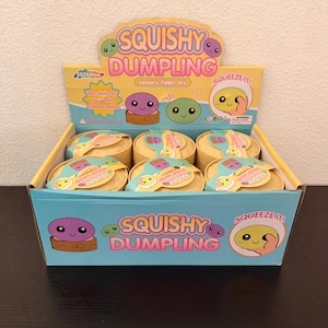 Puede incluir: Una caja expositora llena de juguetes sensoriales Squishy Dumpling. La caja es de color azul claro y amarillo con las palabras "SQUISHY DUMPLING" en rosa y morado. Cada dumpling está en un recipiente amarillo con una cara de dibujos animados.