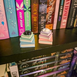 May include: Two miniature stacks of three books each, topped with small animal figurines. One stack features a black cat on light green, mint, and blue books. The other stack features a white cat on lavender, pink, and light pink books.