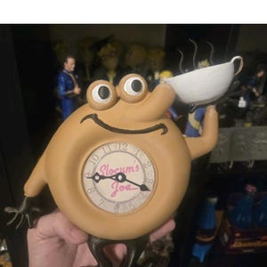 May include: A tan, round, anthropomorphic clock figure with large eyes, a smiling mouth, and dark brown limbs. The clock face features black numbers and the pink text Slocum's Joe. The figure holds a white cup with steam rising from it.