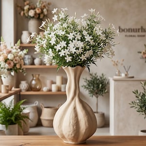 May include: A beige, textured vase with a bulbous base and a slender neck, filled with white jasmine flowers. The vase sits on a wooden table, with other floral arrangements and decor visible in the background, creating a floral shop ambiance.