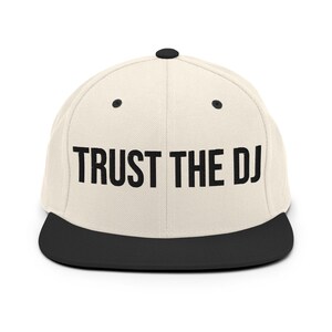 Op de afbeelding: Een crèmekleurige baseballpet met platte klep met de tekst "TRUST THE DJ" in vette zwarte letters. De klep is zwart en de pet heeft een zwarte knoop bovenop. Dit is een stijlvol accessoire voor muziekliefhebbers.