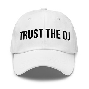 Op de afbeelding: Witte baseballpet met de tekst "TRUST THE DJ" in vette zwarte letters op de voorkant. De pet heeft een gebogen klep en een knoop aan de bovenkant. Een casual accessoire.
