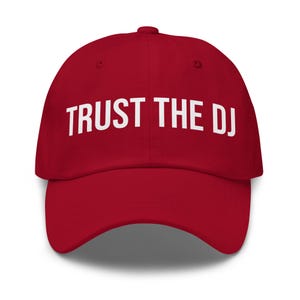 Op de afbeelding: Een rode baseballcap met de witte tekst "TRUST THE DJ" over de voorkant. De cap heeft een gebogen klep en een afgeronde bovenkant. De hoed heeft een effen, levendige rode kleur.