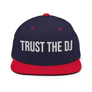Op de afbeelding: Marineblauwe baseballcap met een rode klep en knoop. De witte geborduurde tekst op de voorkant luidt "TRUST THE DJ". De cap heeft rode oogjes. Een modieus accessoire.