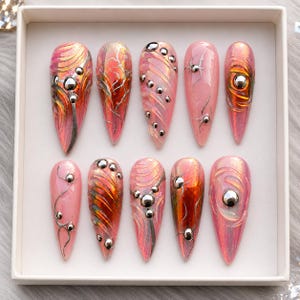 May include: A set of ten almond-shaped press-on nails in a light pink base color with iridescent finishes. The nails feature raised silver metallic accents, swirling textures, and small silver spherical studs arranged in various patterns.