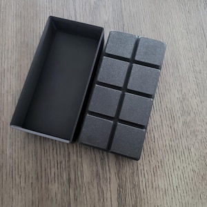 May include: A dark gray rectangular box with a matching lid, next to a dark gray, segmented object. The segmented object is divided into eight square sections. The items are on a wooden surface.