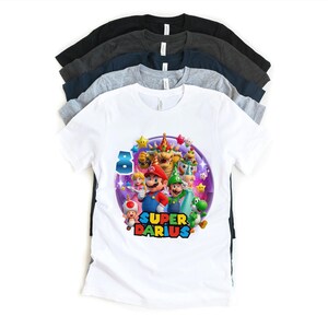 Puede incluir: Una pila de camisetas de manga corta en varios colores, incluyendo blanco, gris, azul marino y negro. La camiseta blanca presenta un gr&aacute;fico colorido de personajes de Super Mario con el texto "SUPER DARIUS" y el n&uacute;mero "8".