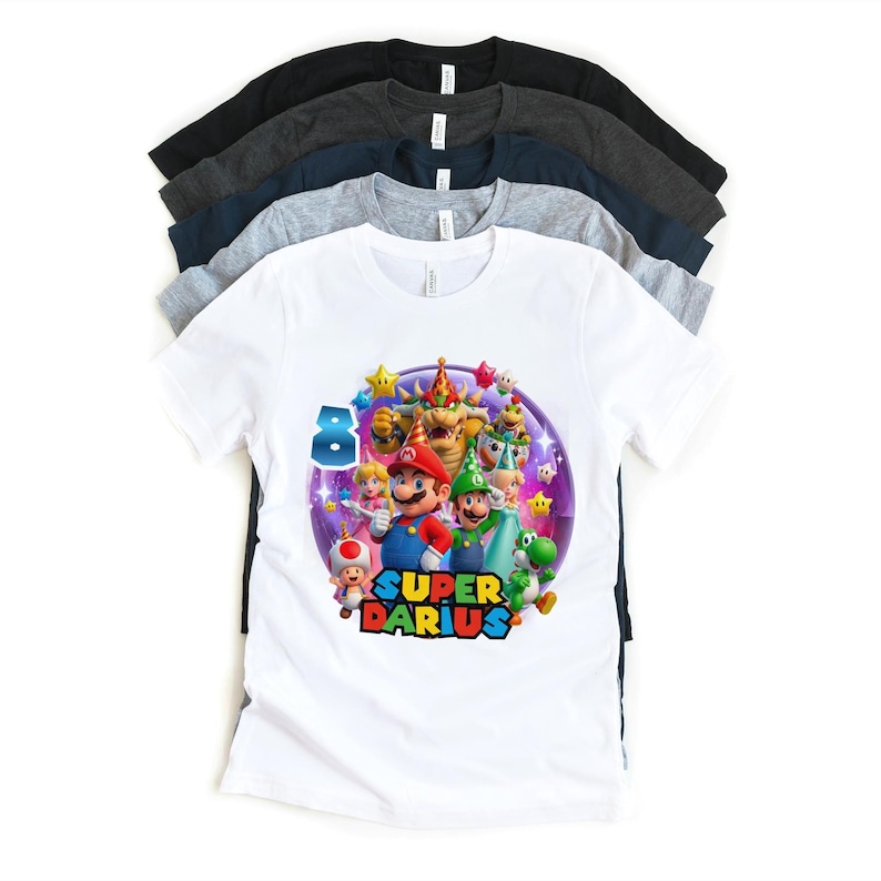 Puede incluir: Una pila de camisetas de manga corta en varios colores, incluyendo blanco, gris, azul marino y negro. La camiseta blanca presenta un gr&aacute;fico colorido de personajes de Super Mario con el texto "SUPER DARIUS" y el n&uacute;mero "8".