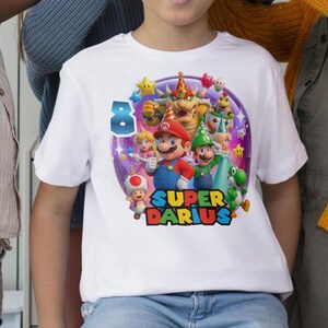 Puede incluir: Camiseta blanca con un gr&aacute;fico colorido de personajes de Super Mario, incluyendo a Mario, Luigi y la Princesa Peach. El dise&ntilde;o incluye el n&uacute;mero 8 y el texto "SUPER DARIUS". La camiseta es de talla infantil.