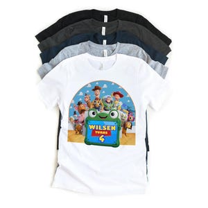 May include: A stack of short-sleeved t-shirts in various colors, including white, gray, and black. The front shirt is white and features a colorful graphic of characters from the movie Toy Story, with the text "Wilsen Turns 4".