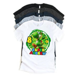 May include: White t-shirt featuring a green Yoshi character with the number 8 and the name "Andrew". The design is set within a green circular frame. Several other t-shirts in various colors are stacked behind it.