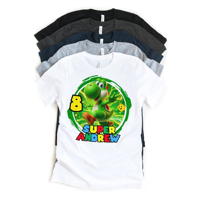 May include: White t-shirt featuring a green Yoshi character with the number 8 and the name "Andrew". The design is set within a green circular frame. Several other t-shirts in various colors are stacked behind it.