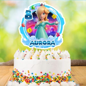 May include: A white frosted cake with colorful sprinkles and a decorative topper. The topper features a cartoon character, the number 8, and the name "Aurora". The character is a blonde woman in a teal dress, surrounded by stars.