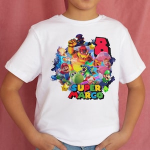 May include: White t-shirt featuring a colorful graphic of Super Mario characters, the number 8, and the text "SUPER MARCO". The design includes Mario, Luigi, Bowser, and other characters from the video game series.