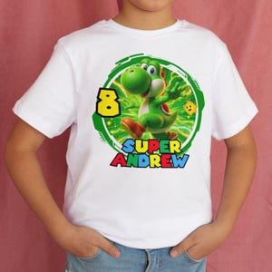 May include: White t-shirt featuring a green Yoshi character from the Super Mario video game series. The design includes the number 8 in yellow, the name "SUPER ANDREW" in red, blue, and yellow, and a green circular border.