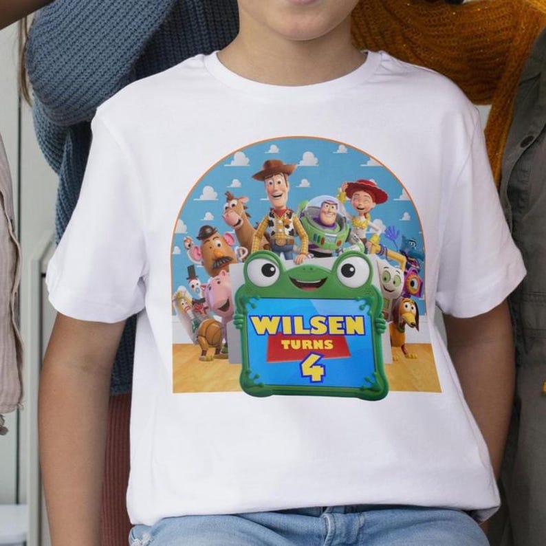 May include: White t-shirt featuring a colorful graphic of characters from the Toy Story movie. The design includes Woody, Buzz Lightyear, and others, with the text "Wilsen Turns 4" on a green frog-shaped sign.