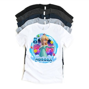 May include: White t-shirt featuring a colorful graphic of a character with blonde hair and a teal dress, surrounded by stars and the name "AURORA". The number "8" is also displayed. Several other t-shirts in various colors are stacked behind it.