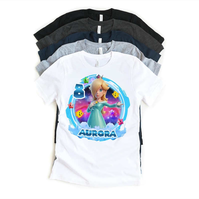 May include: White t-shirt featuring a colorful graphic of a character with blonde hair and a teal dress, surrounded by stars and the name "AURORA". The number "8" is also displayed. Several other t-shirts in various colors are stacked behind it.