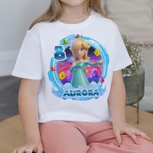 May include: White t-shirt featuring a cartoon graphic of a character in a teal dress with a crown, surrounded by stars and the number 8. The name "AURORA" is printed below the character in blue letters.