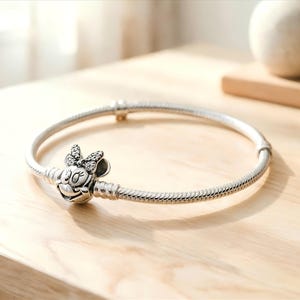 May include: A silver-toned snake chain bracelet featuring a charm shaped like a cartoon mouse head with a bow. The bow is embellished with small, clear stones. The bracelet has a smooth, rounded clasp mechanism and a flexible, textured metal band.