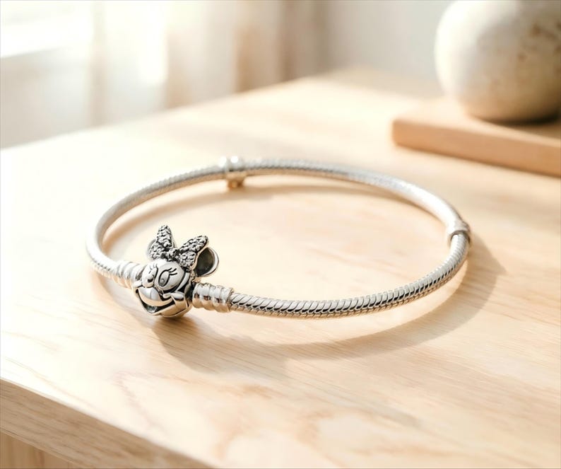 May include: A silver-toned snake chain bracelet featuring a charm shaped like a cartoon mouse head with a bow. The bow is embellished with small, clear stones. The bracelet has a smooth, rounded clasp mechanism and a flexible, textured metal band.