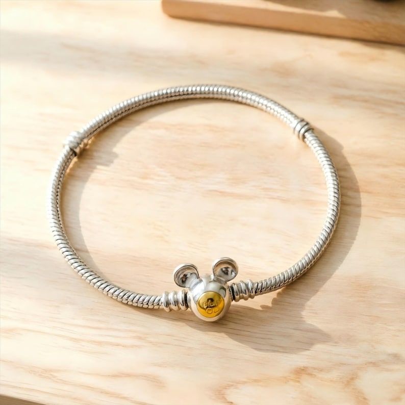 May include: A silver snake chain bracelet featuring a central clasp shaped like a mouse head with two rounded ears and a circular gold-toned accent on the front. The bracelet includes two fixed silver stations along the chain structure.