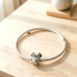 May include: A silver-toned snake chain bracelet featuring a central charm shaped like the head of a cartoon mouse with a bow. The charm is embellished with small clear stones on the bow and has engraved facial features including eyes and a smile.