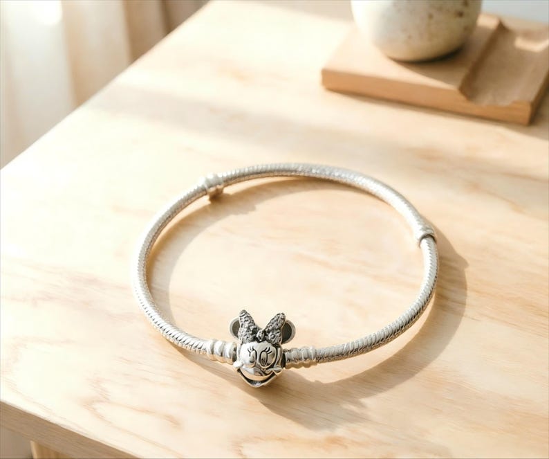 May include: A silver-toned snake chain bracelet featuring a central charm shaped like the head of a cartoon mouse with a bow. The charm is embellished with small clear stones on the bow and has engraved facial features including eyes and a smile.