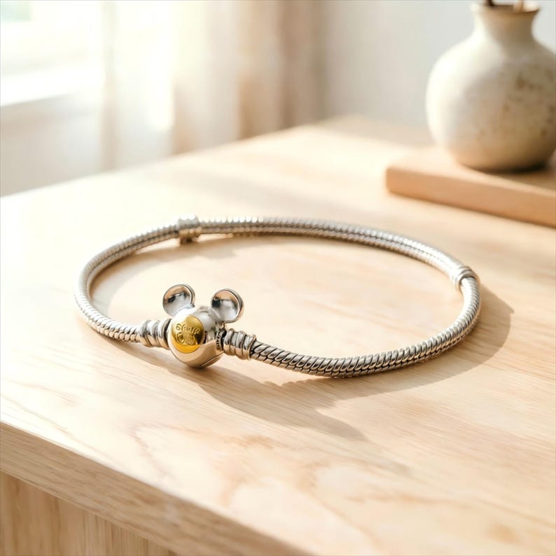 May include: A silver snake chain bracelet featuring a central clasp shaped like a mouse head with two rounded ears. The clasp has a polished silver finish with a circular gold-toned accent in the center, creating a distinct two-tone metallic look.