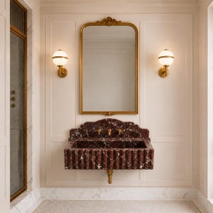 May include: A bathroom interior with a gold-framed mirror above a red marble sink. Two gold sconces with white globe lights are either side of the mirror. The sink has a fluted design and gold fixtures. The walls are a light cream colour.