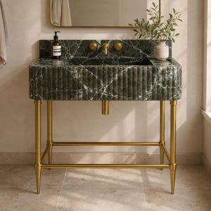 Può includere: Un lavabo da bagno in marmo verde con venature bianche, caratterizzato da un design scanalato. Il lavabo ha rubinetti color oro ed è sostenuto da gambe dorate. Sono visibili uno specchio e un piccolo vaso con verde.
