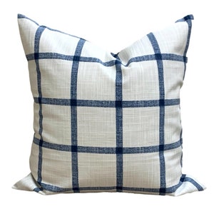 May include: Square cushion featuring a white backdrop with a navy blue grid pattern. The cushion has a textured finish and is intended for decorative use on a bed or sofa.
