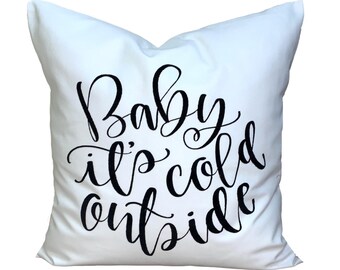 May include: A white square pillow with the phrase "Baby it's cold outside" in black cursive script. The pillow is a decorative item, likely for a bed or couch, and evokes a winter theme.